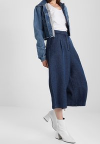 Jeansowa kurtka nałożona na białą koszulkę, w połączeniu z szerokimi, wysokimi niebieskimi spodniami dżinsowymi i błyszczącymi białymi botkami za kostkę.