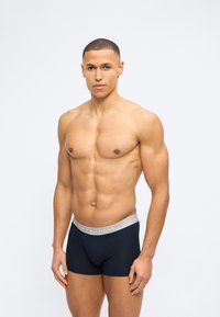 Μυώδης άνδρας με κοντά μαλλιά που φοράει σκούρα μπλε ανδρικά εσώρουχα Tommy Hilfiger σε απλό λευκό φόντο.