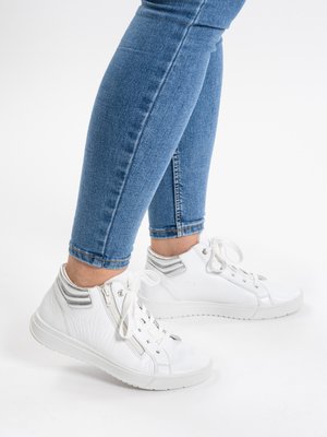Person iført blå jeans og hvide høje sneakers med snørebånd og sidelomme lynlås, stående mod hvid baggrund.