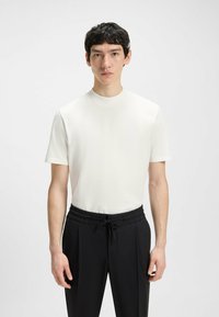 Weißes Kurzarmhemd mit hohem Kragen, kombiniert mit schwarzen Hosen mit elastischem Bund und Kordelzug. Glatte Textur.