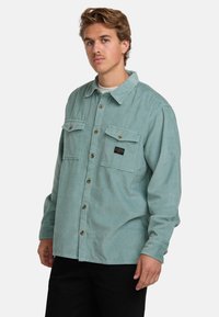 Lys teal kordfløjl skjorte med knaplukning foran, to brystlommer og et sort logo-patch. Har krave og lange ærmer.