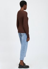 Eine Person steht seitlich und trägt einen braunen strukturierten Pullover, hellblaue, hochgekrempelte Jeans und schwarze Loafers mit goldener Kettendetail.