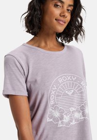 Camiseta de manga corta en color lavanda, fabricada en un material suave, que presenta un diseño gráfico blanco con un sol y acentos florales en la parte inferior.
