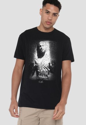 Young man wearing a black Star Wars t-shirt with a black-and-white image of a character on the front, paired with beige pants.