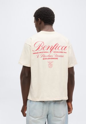 Νέος άνδρας με κοντά κατσαρά μαλλιά φοράει μπεζ μπλουζάκι με κόκκινο κείμενο "Benfica" και έμβλημα, σε συνδυασμό με ανοιχτό μπλε τζιν, με την πλάτη στην κάμερα.