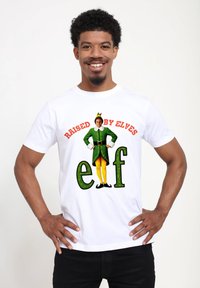 Biała bawełniana koszulka z nadrukiem przedstawiającym tekst "Wychowany przez Elfy" nad postacią elfa w zielonym ubraniu i żółtych nogawicach.