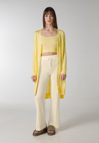 Geel vest over een beige crop top en beige wijde broek met een trekkoord in de taille. Bruine slide sandalen maken de outfit compleet.