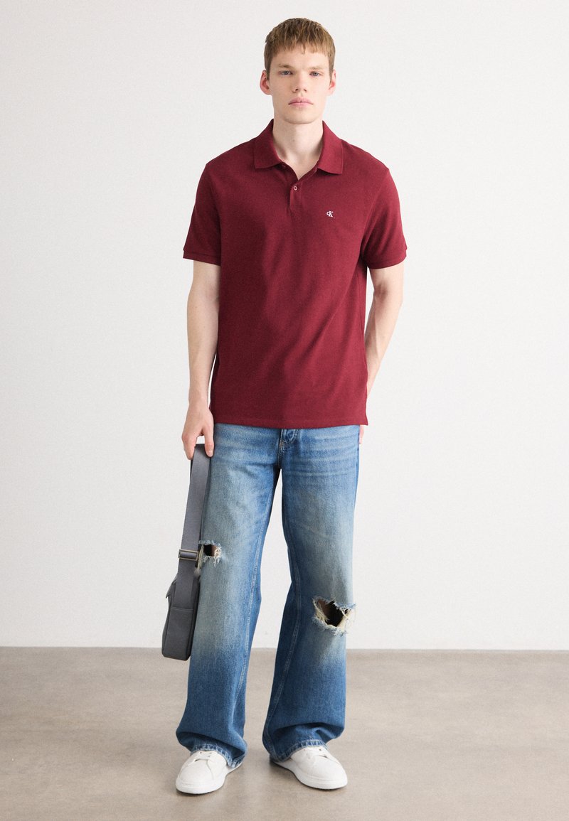 Bordowa koszulka polo z kołnierzykiem, w połączeniu z jasnoniebieskimi dżinsami z przetarciami i dużymi dziurami. Model trzyma szary plecak i ma na sobie białe sneakersy.