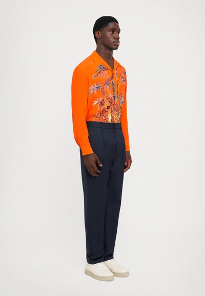 Lys orange skjorte med blomster- og tigermønster, lange ærmer og krave, parret med mørkeblå bukser og hvide sko.