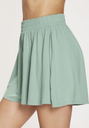 Frau trägt einen hellgrünen, fließenden Rock mit elastischem Bund, der oberhalb der Knie endet, vor einfarbigem Hintergrund.