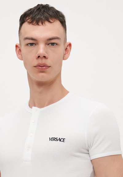 Weißes, geripptes Poloshirt mit kurzen Ärmeln, das über eine klassische Knopfleiste und ein schwarzes, gesticktes "VERSACE"-Logo auf der linken Brust verfügt.