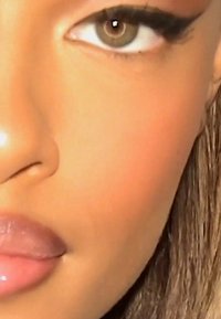 Gros plan sur un visage montrant un œil vert-brun avec un eye-liner défini, une peau lisse de couleur marron clair et des lèvres roses pulpeuses. Les cheveux sont raides et clairs.