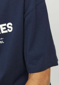 Camiseta de algodón azul marino con cuello redondo, mangas cortas y texto impreso en blanco. La tela es suave y tiene un ligero brillo.