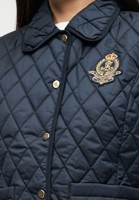 Μπλε τζάκετ καπιτονέ με κλασικό γιακά, κλεισίματα με χρυσά κουμπιά και κεντημένο λογότυπο στην τσέπη του στήθους.