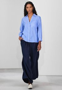 Light blue striped button-up shirt with a chest pocket, paired with loose navy pants and white slip-on shoes, displayed against a gray background.