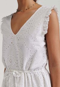 White cotton dress featuring a V-neckline, ruffled sleeves, intricate broderie anglaise pattern, and adjustable drawstring waist.