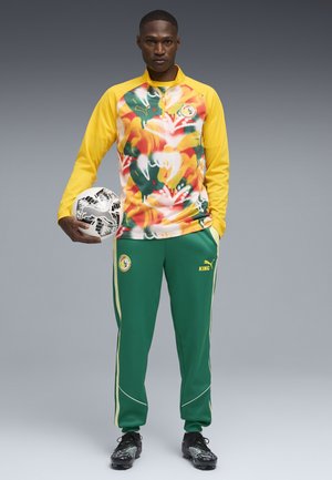 Mann in gelbem und mehrfarbigem floralem Sportoberteil und grüner Hose hält einen schwarz-weißen Fußball und trägt schwarze Fußballschuhe vor grauem Hintergrund.