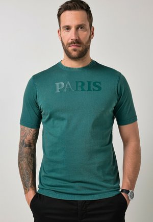 Hombre con barba y brazo izquierdo tatuado, vistiendo una camiseta verde azulado con la palabra "PARIS" y pantalones negros, con las manos en los bolsillos, fondo neutro.