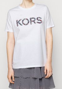 Bílý bavlněný tričko s nápisem "KORS" v květinovém vzoru. Krátké rukávy, kulatý výstřih, v kombinaci s sukni s volánovým vzorem.