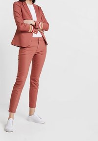 Rosa kavaj med guld-accenterade manschetter, matchad med figursydda byxor och vita sneakers. Slätt tyg, skräddarsydd design och elegant silhuett.