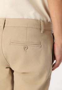 Beige pamut nadrág hátul kialakított zsebével, amely gombbal záródik. Az anyag sima tapintású, és a dizájn övhurkokkal rendelkezik.