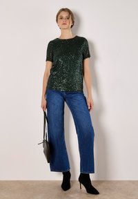 A woman stands confidently wearing a glittery green top, blue wide-leg jeans, and black heeled boots, holding a small black bag.