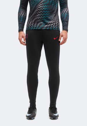 Bărbat purtând pantaloni sport negri, comoți, cu sigle roșii Nike și PSG, ghete negre și un top cu mâneci lungi și model zigzag.