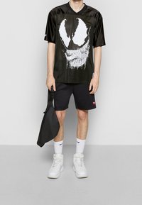 Black mesh jersey with a large white graphic design featuring a skull and fangs, paired with black shorts and white sneakers. Black bag held.