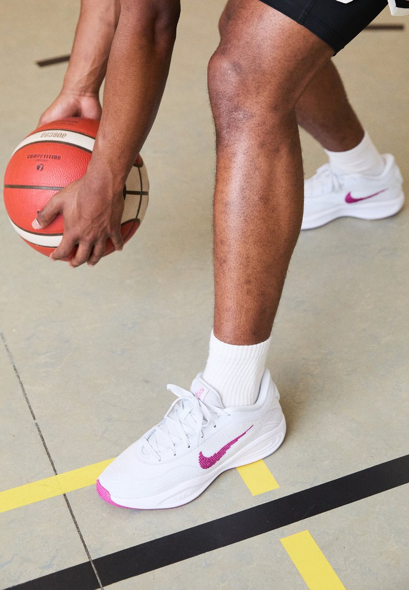 Des chaussures de basket Nike blanches avec des accents violets, dotées d'un dessus texturé et d'une semelle épaisse, associées à des chaussettes blanches et des shorts noirs.