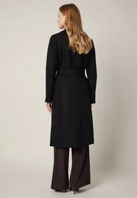 Czarny długi płaszcz z paskiem w talii, dopasowany fason i wyraźnie zarysowane ramiona, w komplecie z brązowymi spodniami o szerokich nogawkach w prążki.