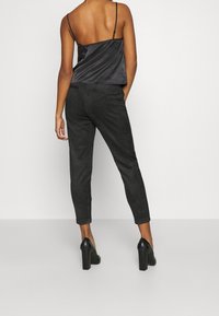 Haut caraco en satin noir à fines bretelles, associé à un pantalon taille haute foncé texturé et des chaussures noires à talons blocs.