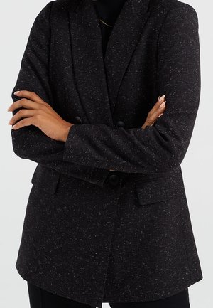 Persoon met gekruiste armen, gekleed in een zwart gemêleerd colbert over een zwarte top, zichtbaar van schouders tot heupen tegen een effen achtergrond.