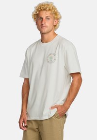 Camiseta de algodón blanca con un parche de logo redondo en verde y negro en el lado izquierdo del pecho. Mangas cortas, corte relajado, y dobladillo ligeramente curvado.