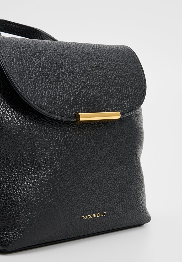 Czarna skórzana torebka o fakturowanym wykończeniu, z klapą i złotym elementem ozdobnym. Nazwa marki "COCCINELLE" wytłoczona na złoto.