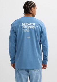 Camisa de algodón azul de manga larga con texto blanco en la parte posterior, incluyendo "MIDWEST STUDIOS" y detalles de dirección. Textura suave, corte relajado.