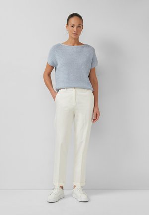 Frau steht und trägt ein hellblaues strukturiertes kurzärmeliges Oberteil, weiße hoch taillierte Hose, weiße Sneakers und Creolen vor einem grauen Hintergrund.