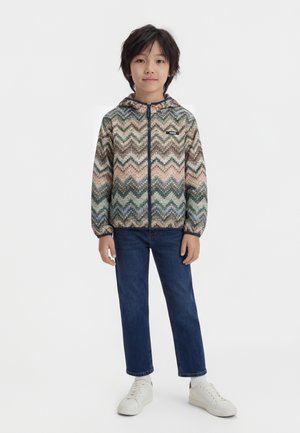 Kind steht frontal und trägt einen mehrfarbigen, chevronmusternden Kapuzenpullover mit Reißverschluss, blaue Jeans, weiße Socken und weiße Turnschuhe.