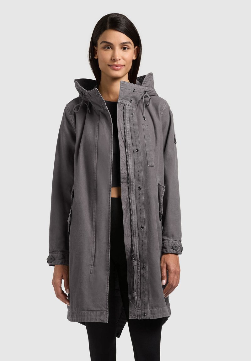 Donna che indossa un lungo cappotto grigio con cappuccio, con cerniera frontale e bottoni a pressione, sopra un top nero cortissimo e pantaloni neri, in piedi davanti a uno sfondo semplice.