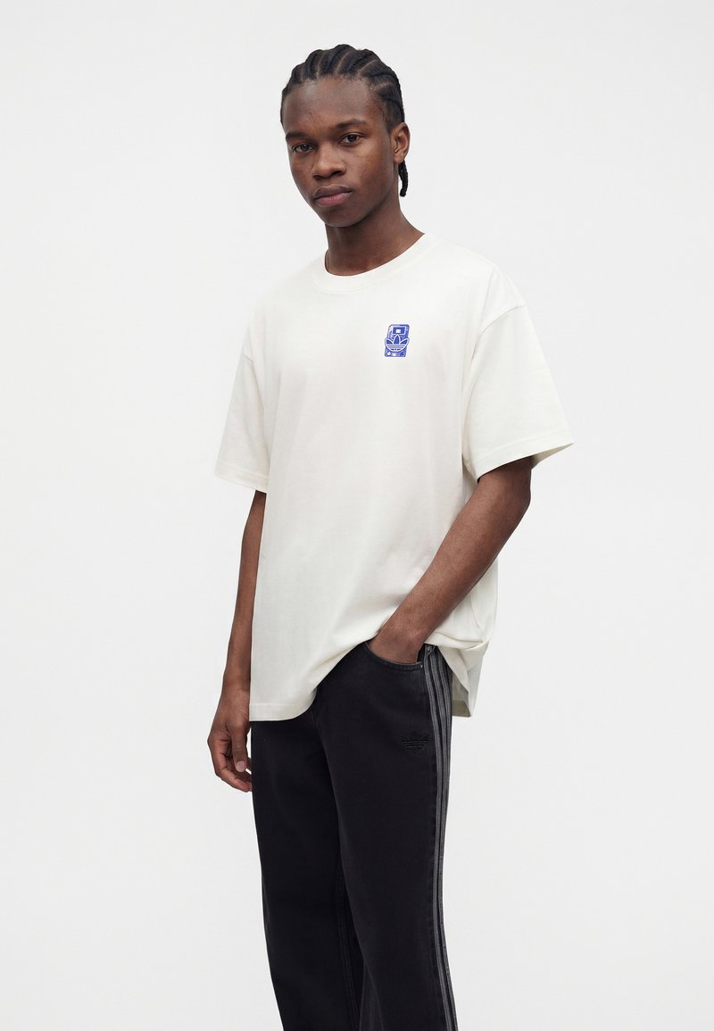 Jeune homme aux cheveux tressés portant un t-shirt blanc avec un motif bleu et un pantalon noir avec des bandes latérales, debout avec une main dans la poche.