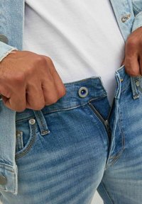 Ljusblå denim jeans med dragkedja och knäppning, med synlig sömnad och en lätt blekning. Hand som håller i midjebandet.