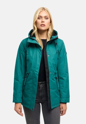 Femme portant une veste à capuche bleu sarcelle sur un haut noir et un jean gris, debout avec une expression neutre sur un fond uni.