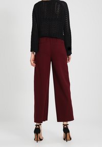Persona che indossa pantaloni larghi color bordeaux e un top a maglia lungo nero, in piedi con la schiena rivolta verso l'osservatore su uno sfondo semplice.