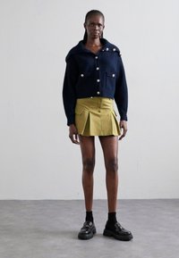 Navy knitted cardigan with large buttons and a hood, paired with a pleated lime green skirt. Model wears black chunky platform shoes.