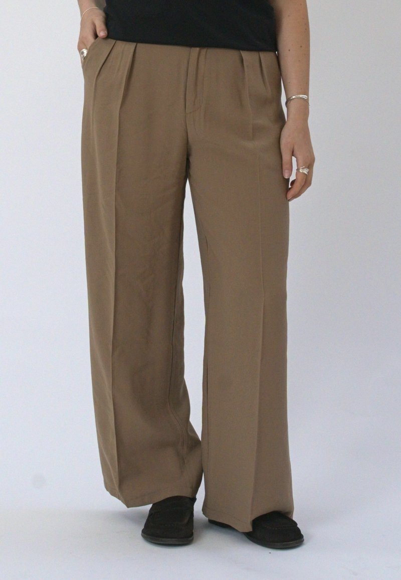 Personne portant un pantalon marron ample, des chaussures noires, un haut noir, avec la main dans la poche et portant des bagues et des bracelets.