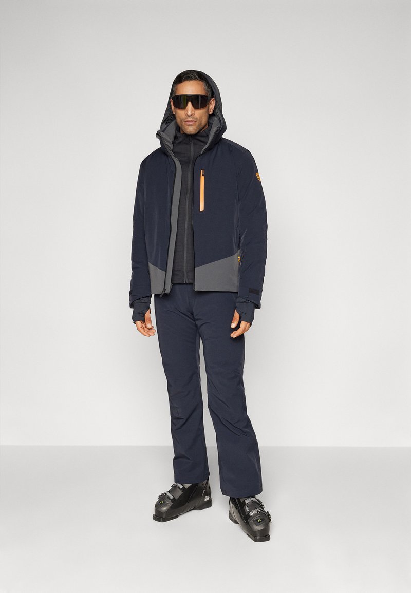 Giacca outdoor blu navy e grigia con cerniere arancioni, abbinata a pantaloni coordinati. Il modello indossa occhiali da sole e scarponi da sci.