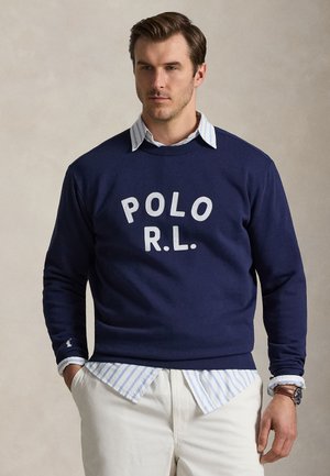 Homme portant un sweat-shirt bleu marine avec le texte "POLO R.L.", sur une chemise rayée bleue à boutons, associé à un pantalon blanc et une montre-bracelet.