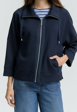Donna che indossa una giacca blu navy con zip sopra una camicia a righe blu e jeans blu, con una mano nella tasca della giacca.