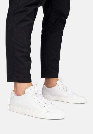 Persona che indossa scarpe da ginnastica bianche basse con lacci e pantaloni neri corti su uno sfondo chiaro.