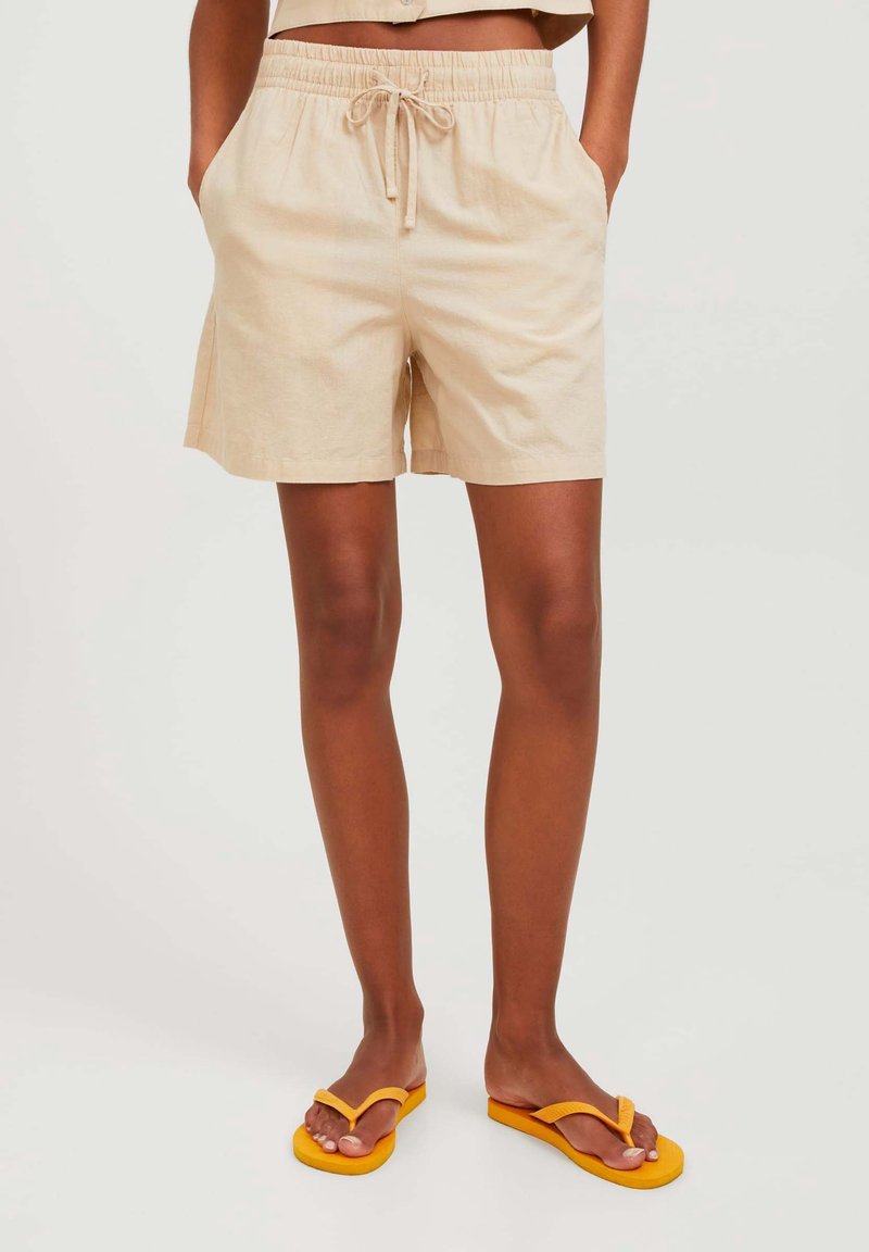 Beige linneshorts med elastisk midjeband och dragsko, med sidofickor. Bärs med gula flip-flops. Slät textur.
