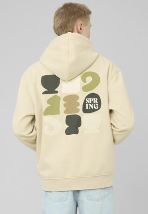Personne portant un sweat à capuche beige avec des formes abstraites et le texte « SPRING » dans le dos, associée à un jean bleu clair, vue de dos.